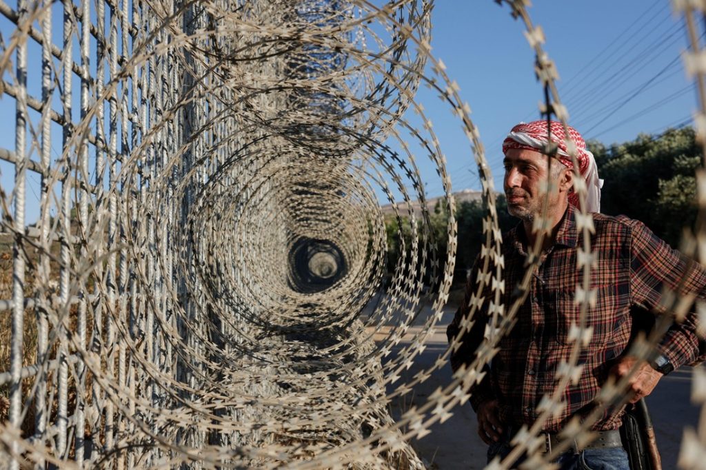 A man wearing a checkered shirt and a red-and-white headscarf standing beside a fence topped with concentric coils of razor wire.