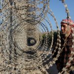 A man wearing a checkered shirt and a red-and-white headscarf standing beside a fence topped with concentric coils of razor wire.