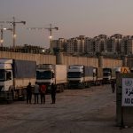 Humanitarian aid trucks parked along a concrete border wall with watchtowers at dusk, next to a sign reading "Aid Distribution Point - Gaza", while apartment buildings and construction cranes rise in the background.