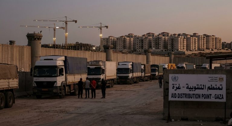 Humanitarian aid trucks parked along a concrete border wall with watchtowers at dusk, next to a sign reading "Aid Distribution Point - Gaza", while apartment buildings and construction cranes rise in the background.