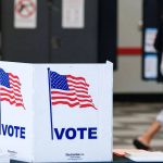 A close-up of a white voting booth featuring an American flag design and a "VOTE" sign inside a polling station.