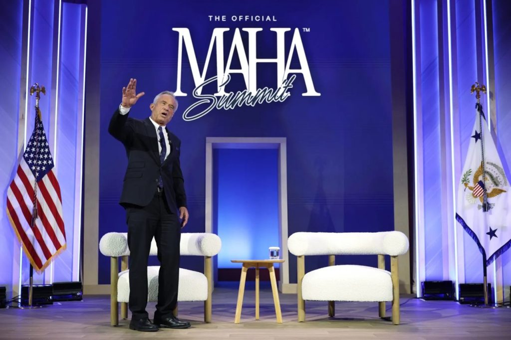 Robert F. Kennedy Jr. waving to an audience on stage in front of a Make America Healthy Again banner.