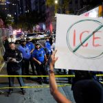 Protester holding a crossed-out ICE sign in front of a police line on a city street.