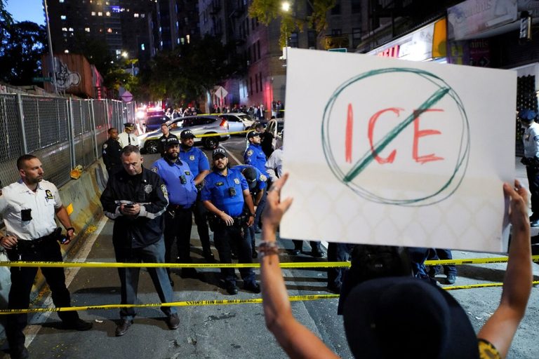 Protester holding a crossed-out ICE sign in front of a police line on a city street.