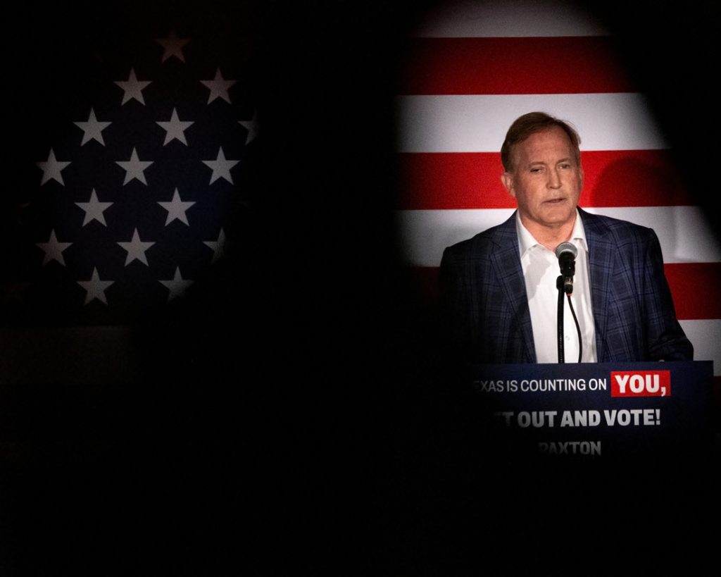 Ken Paxton speaking into a microphone at a podium in front of a dimly lit American flag, with a sign reading Texas is counting on you, get out and vote.