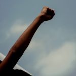 Low-angle shot of a protester's raised arm and clenched fist silhouetted against a cloudy blue sky.