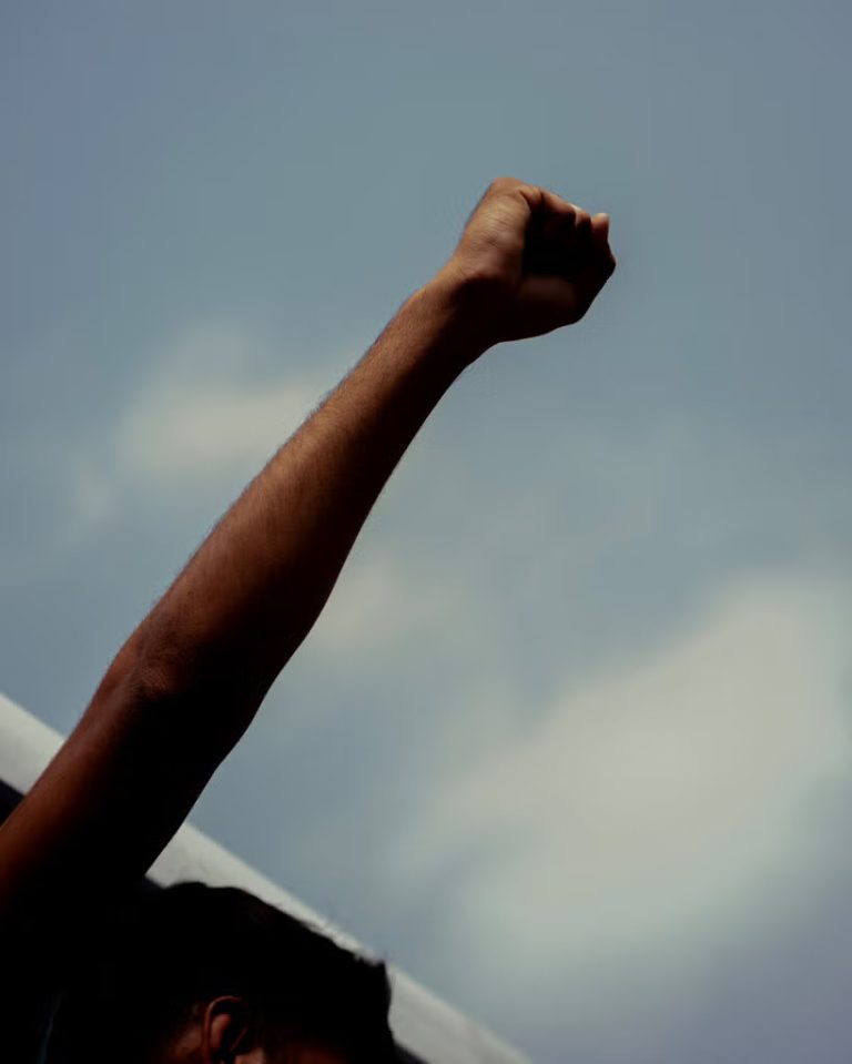 Low-angle shot of a protester's raised arm and clenched fist silhouetted against a cloudy blue sky.