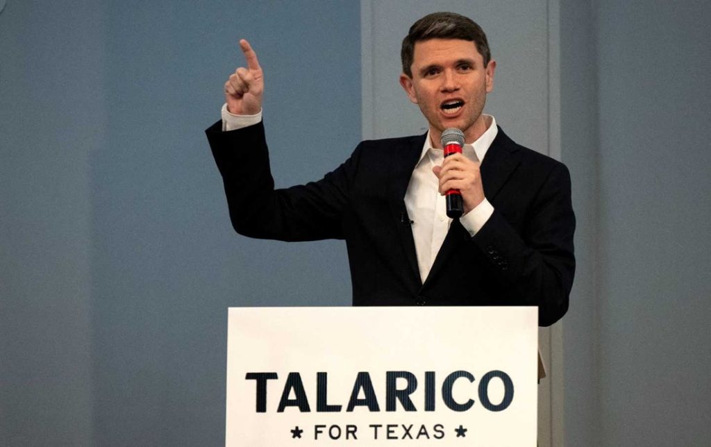 Democratic politician James Talarico speaking into a microphone and pointing his right index finger upward behind a podium that reads "Talarico For Texas".