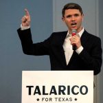 Democratic politician James Talarico speaking into a microphone and pointing his right index finger upward behind a podium that reads "Talarico For Texas".