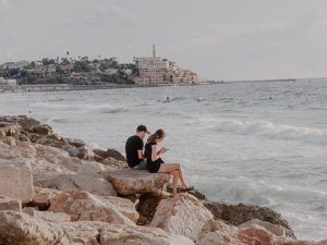 A young couple sitting on coastal rocks looking toward the Mediterranean Sea with Old Jaffa in the background.