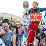 Protester atop a stepladder holding a large caricature puppet and an Israeli flag among a crowd.