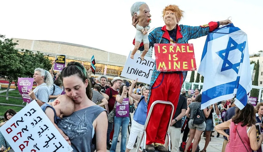 Protester atop a stepladder holding a large caricature puppet and an Israeli flag among a crowd.