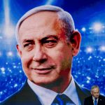 Benjamin Netanyahu standing below a large digital screen displaying a close-up projection of his face.