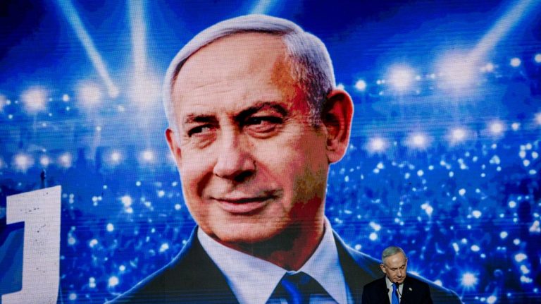 Benjamin Netanyahu standing below a large digital screen displaying a close-up projection of his face.