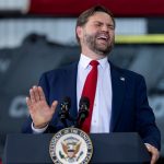 JD Vance speaking behind a presidential podium with a red tie and an expressive gesture.