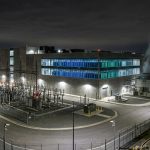A massive industrial data center building at night with illuminated server rooms, outdoor cooling infrastructure, and security fencing.