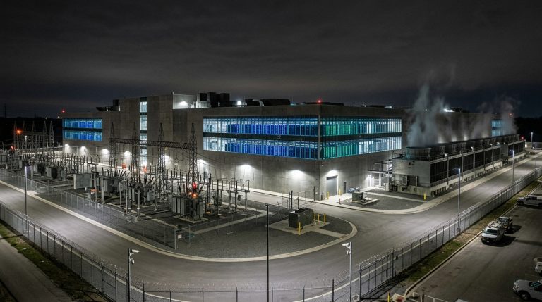 A massive industrial data center building at night with illuminated server rooms, outdoor cooling infrastructure, and security fencing.