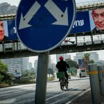 A cyclist riding down an urban street in Israel beneath an overpass billboard showing portraits of Gadi Eisenkot and Benjamin Netanyahu, framed by a blue dual-arrow traffic sign.