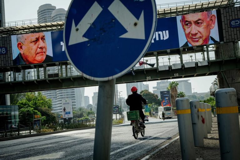 A cyclist riding down an urban street in Israel beneath an overpass billboard showing portraits of Gadi Eisenkot and Benjamin Netanyahu, framed by a blue dual-arrow traffic sign.