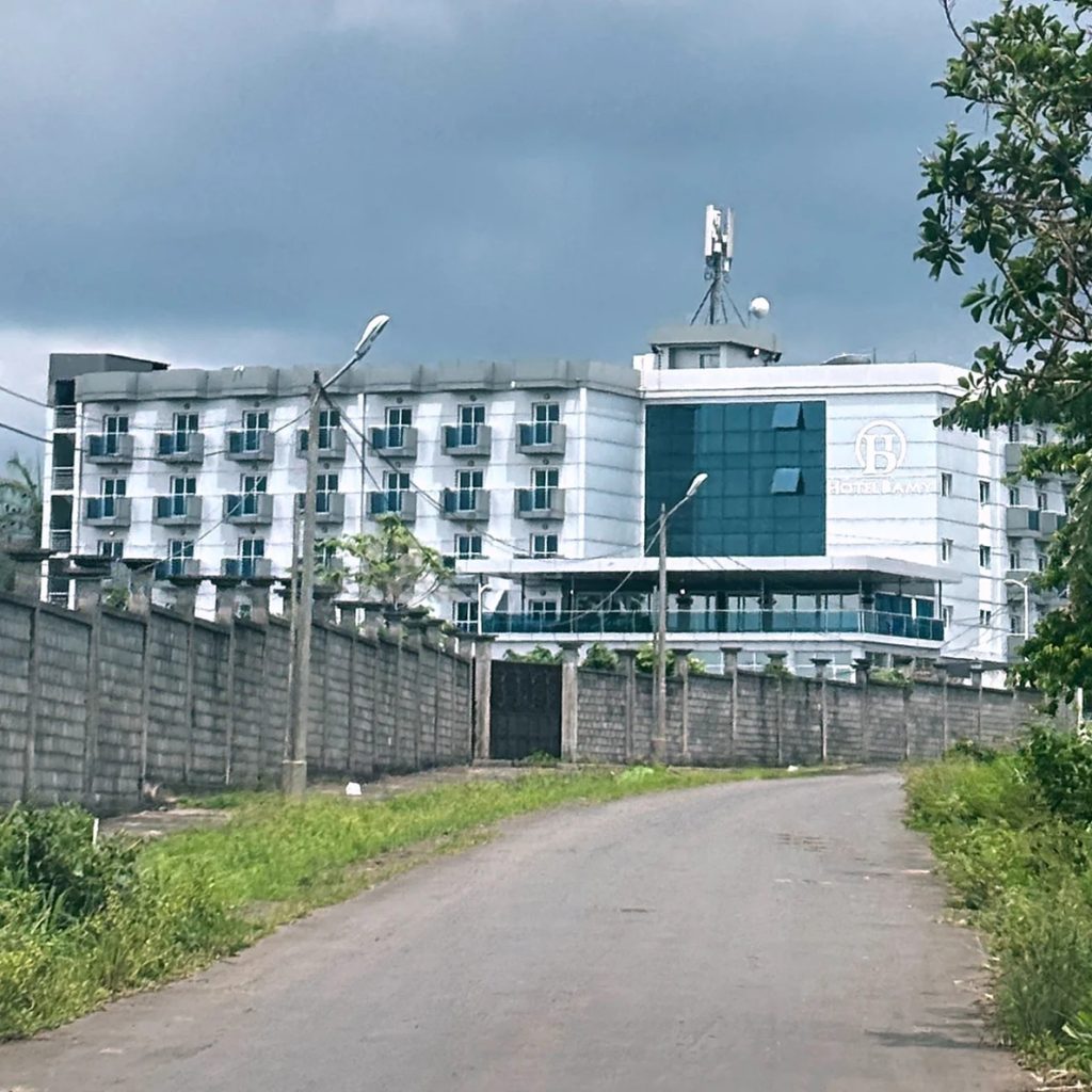 Multi-story white and blue hotel building bordered by a tall concrete security wall along a rural road.