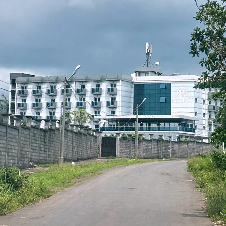 Multi-story white and blue hotel building bordered by a tall concrete security wall along a rural road.