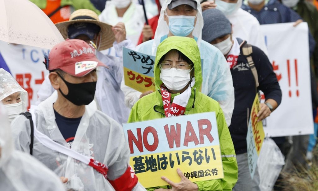 Japanese demonstrator wearing a raincoat and face mask holding a sign that reads No War in Okinawa.