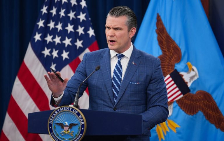 Pete Hegseth speaking at a podium bearing the Department of War seal in front of American and defense flags.