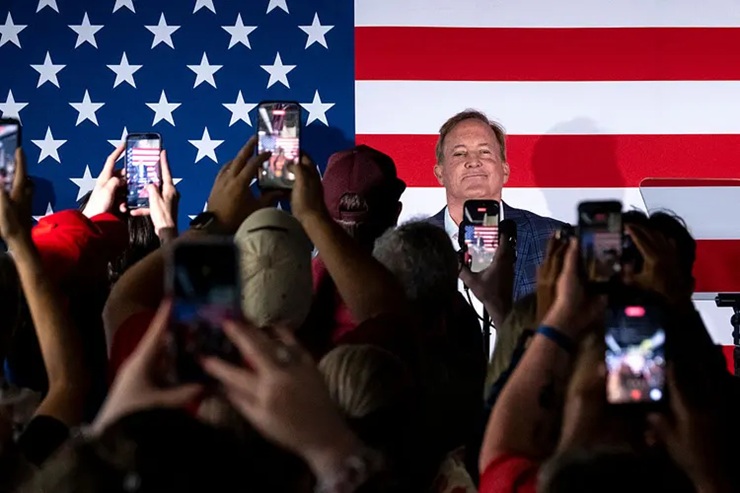 A crowd of supporters holding up their smartphones to record a speaker standing in front of a large American flag.
