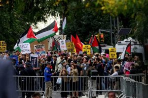 Protesters holding Palestinian flags and demonstration signs behind metal police barricades.