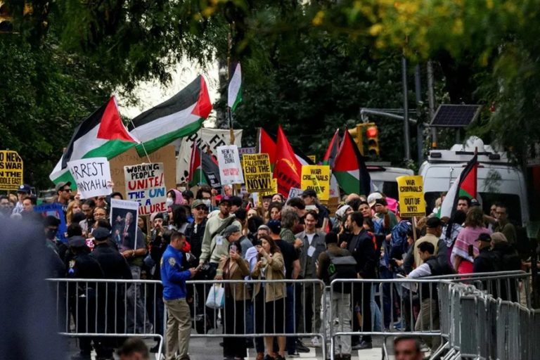 Protesters holding Palestinian flags and demonstration signs behind metal police barricades.