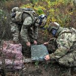 Two soldiers in camouflage uniforms and tactical gear inspecting a Starlink satellite receiver terminal on the ground.
