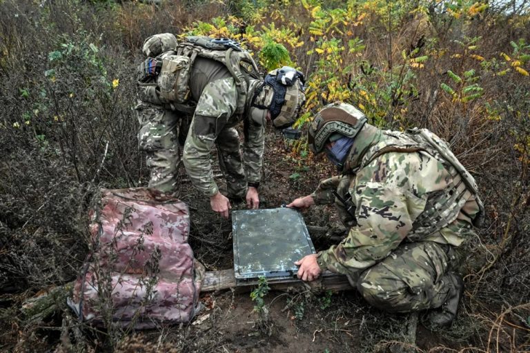 Two soldiers in camouflage uniforms and tactical gear inspecting a Starlink satellite receiver terminal on the ground.
