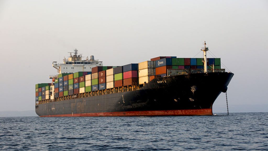 A large black cargo container ship stacked with multicolored shipping containers anchored at sea.