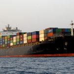 A large black cargo container ship stacked with multicolored shipping containers anchored at sea.