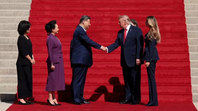 Donald Trump shaking hands with Xi Jinping on a red-carpeted staircase with Melania Trump and Peng Liyuan