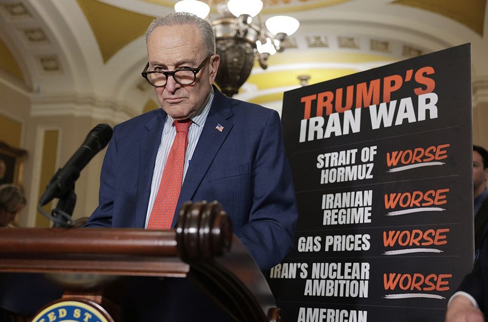 Senator Chuck Schumer speaking at a podium next to a chart criticizing Trump's Iran policy