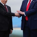 Vladimir Putin in a dark suit and red tie shaking hands with Donald Trump in a navy suit and bright red tie in front of an airplane.