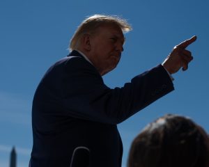Silhouette of Donald Trump pointing his right index finger upward against a blue sky.