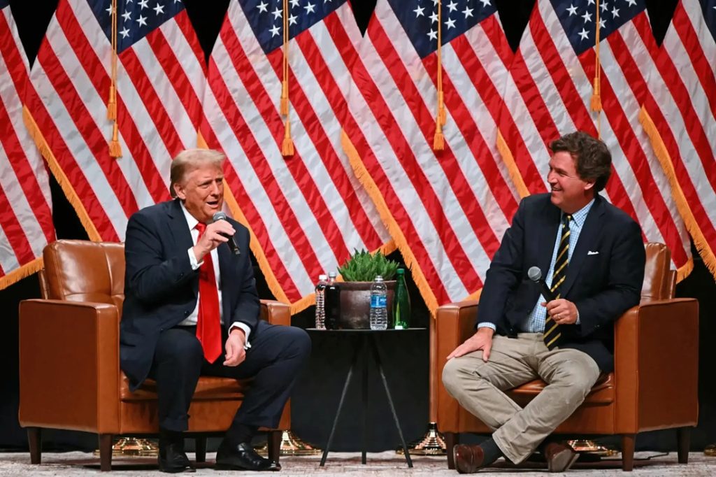 Donald Trump speaking into a microphone seated across from Tucker Carlson on stage in front of American flags.