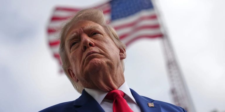 Low-angle portrait of Donald Trump with an American flag waving in the background.