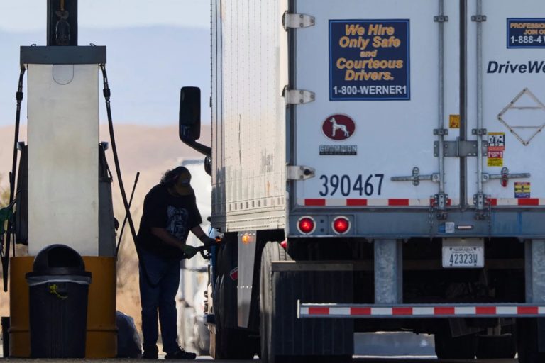A trucker pumping diesel fuel into a large commercial semi-truck at a fueling station.