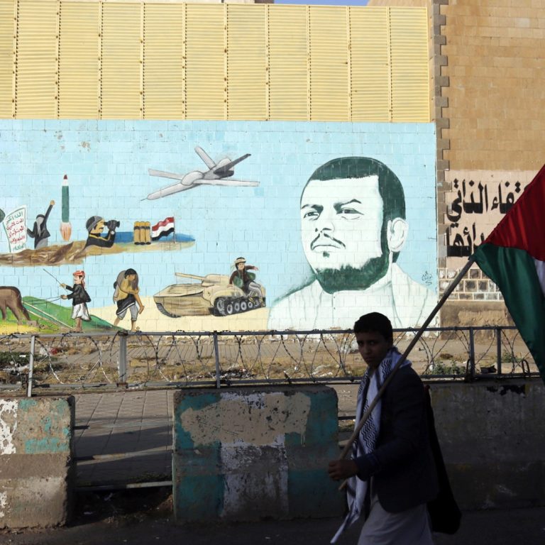 A young person carrying a Palestinian flag past a street mural featuring a military figure, drone, and combat imagery.
