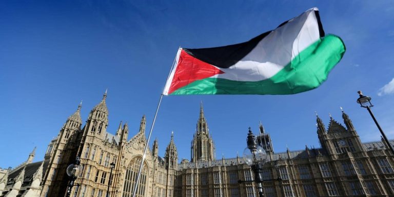 The Palestinian flag waving against a clear blue sky in front of the historic Palace of Westminster in London.