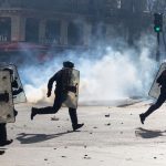 Riot police in tactical gear running through a smoke-filled street during clashes.