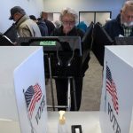 Voters standing at private electronic voting booths with American flag "VOTE" decals on the privacy screens.