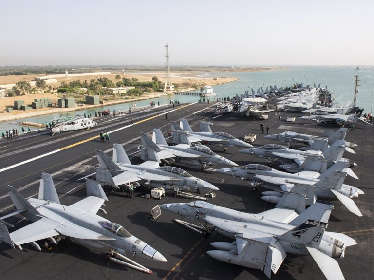 Flight deck of a US Navy aircraft carrier filled with fighter jets navigating through a canal.